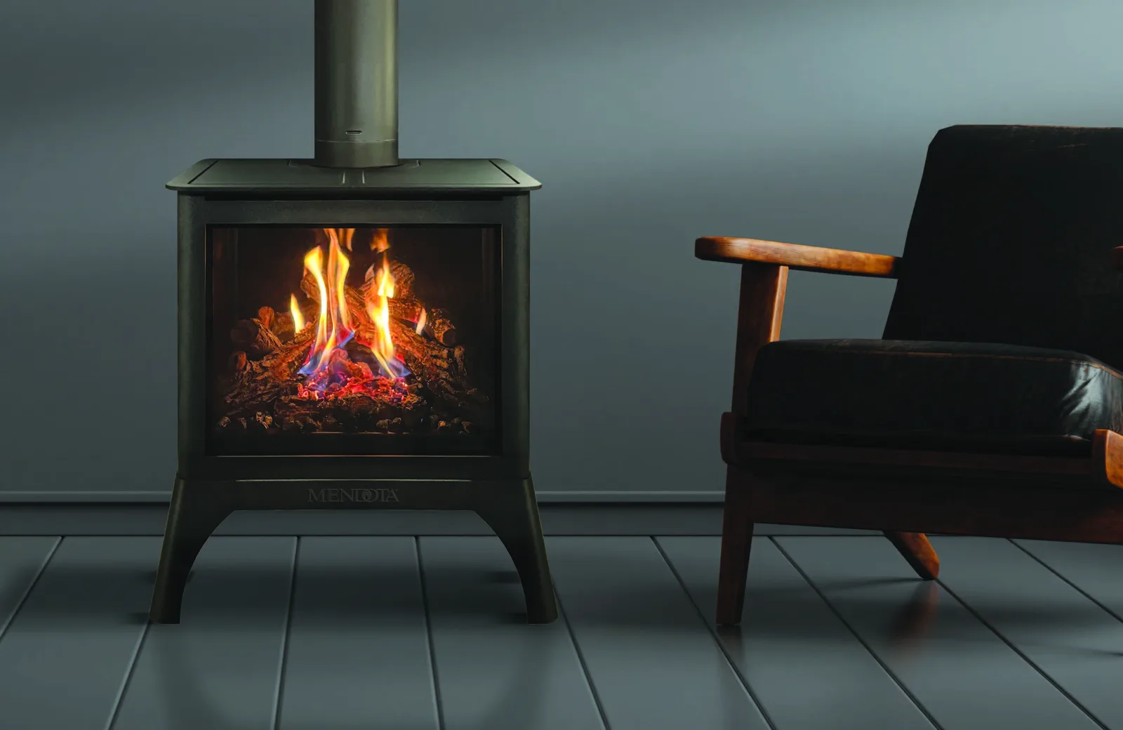 A Mendota 360 FireIn gas stove burning beside a reading chair against a deep teal wall