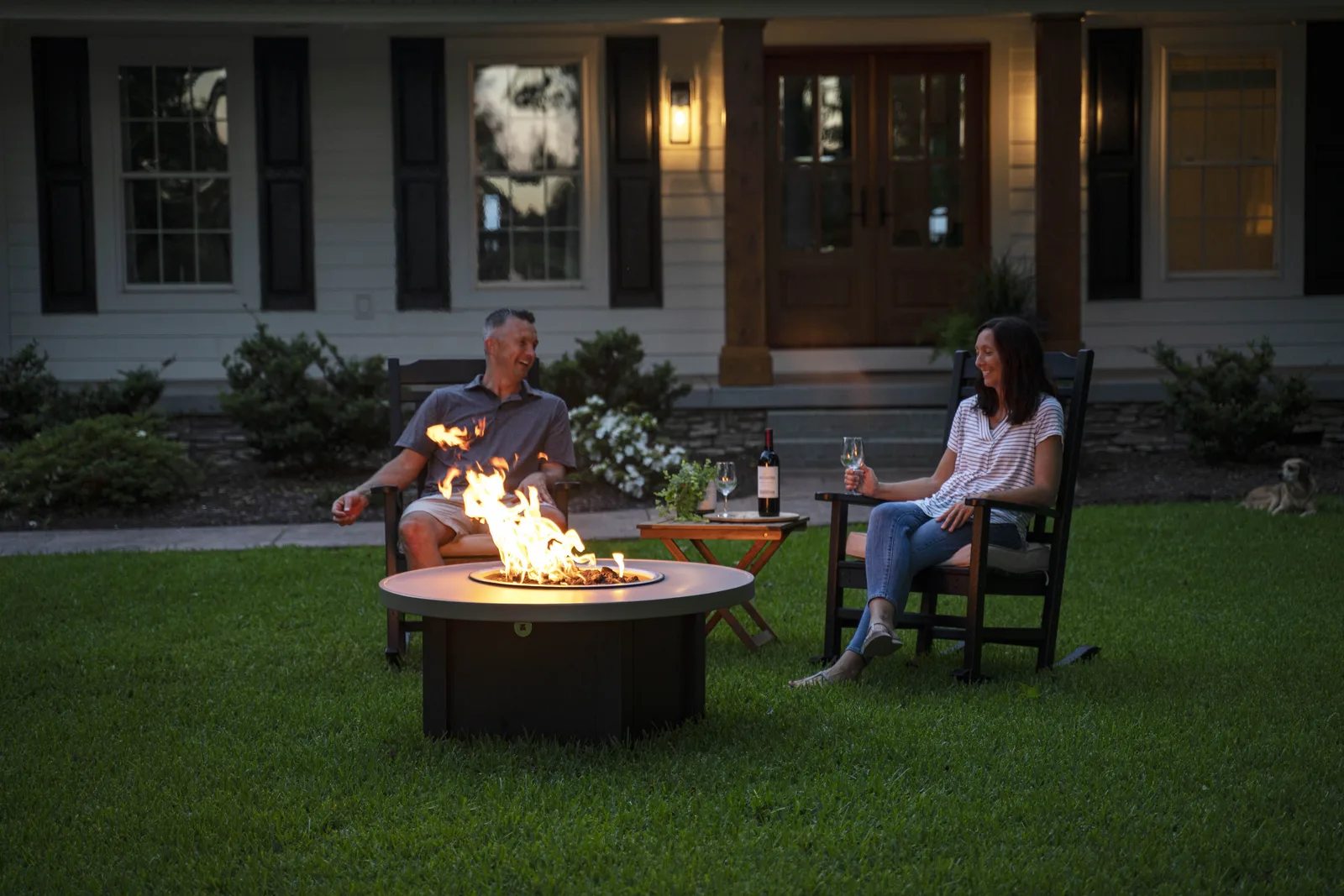 A couple relaxing in rocking chairs beside a round gas fire pit table at dusk, with the front porch lit behind them