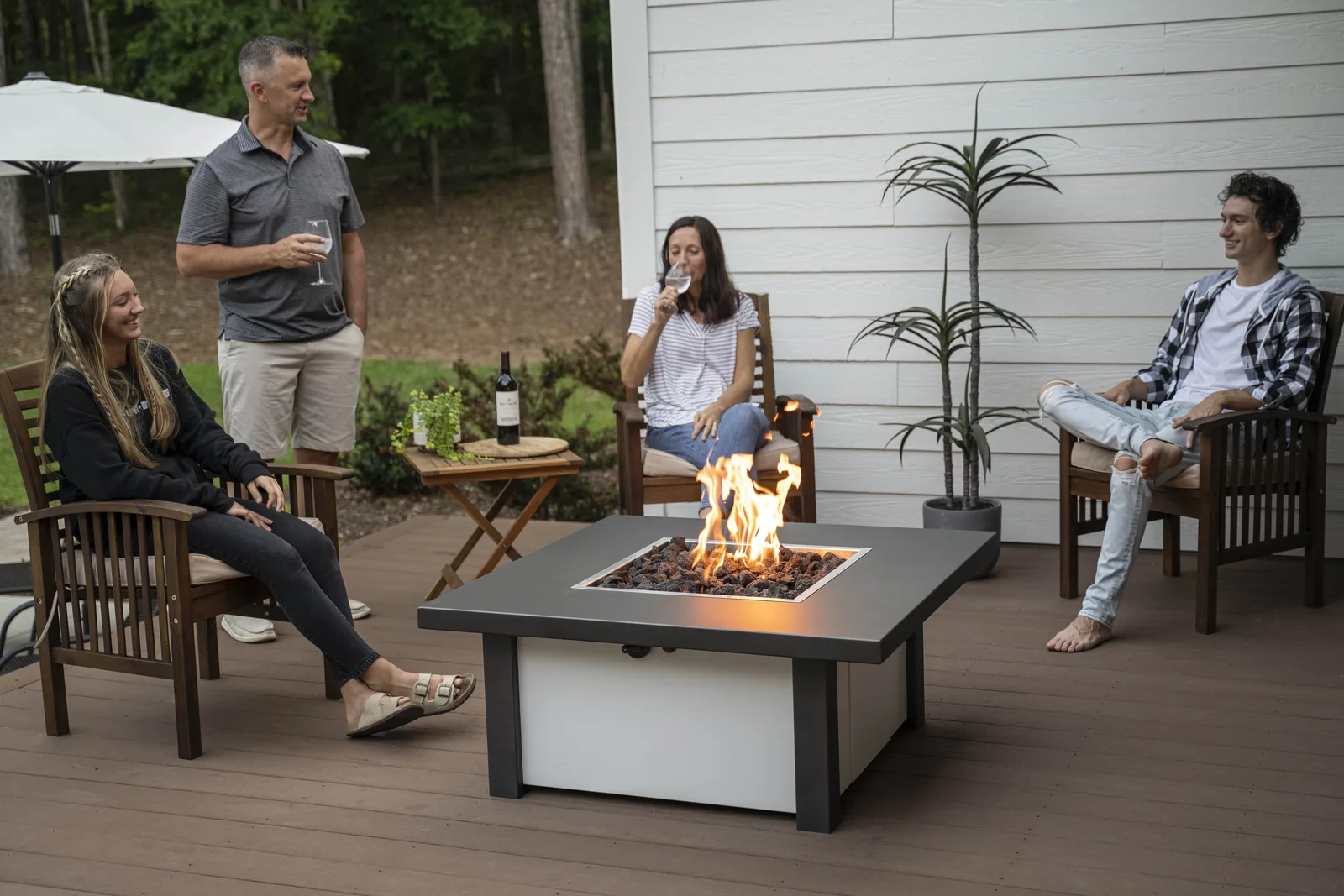 Four people gathered around a lit square gas fire pit table on a backyard deck