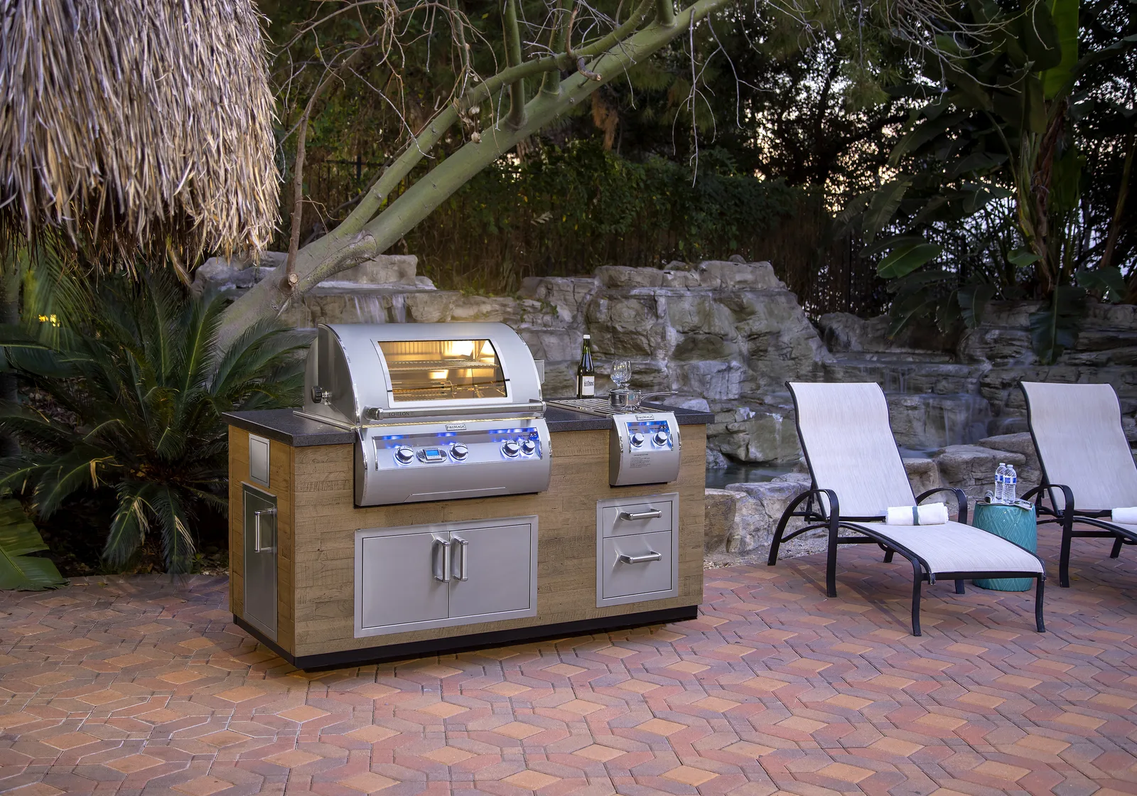 A lit Fire Magic Echelon grill in a wood-paneled island with stainless doors and drawers on a poolside patio at dusk