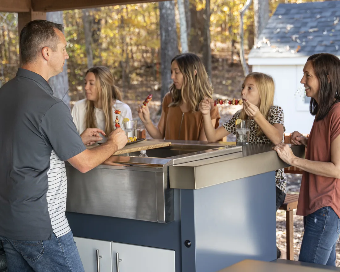 A family gathered around a built-in gas grill in a finished stone outdoor kitchen at golden hour